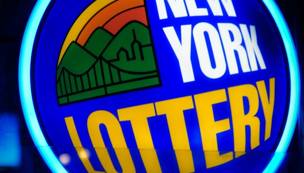 A bright, circular neon sign with a blue background, featuring the words 'New York Lottery' in bold white and yellow letters. The sign includes a colorful illustration of a landscape with green mountains, an orange-red sunset, and a bridge, all encapsulated within the circle.