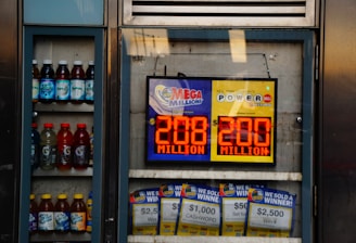 An advertisement display in a store window features lottery jackpot amounts for Mega Millions and Powerball, both showing jackpots of 208 million and 200 million respectively. Below the display, several lottery tickets and promotional posters are visible. To the left of the display, there is a refrigerator stocked with various drinks including different flavors of sports drinks and bottled water.