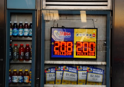 An advertisement display in a store window features lottery jackpot amounts for Mega Millions and Powerball, both showing jackpots of 208 million and 200 million respectively. Below the display, several lottery tickets and promotional posters are visible. To the left of the display, there is a refrigerator stocked with various drinks including different flavors of sports drinks and bottled water.