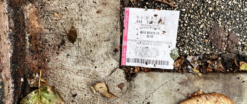 A lottery ticket is lying on a textured concrete surface beside fallen leaves, suggesting it is outdoors. The ground is a mix of dark, wet patches and dry, cracked cement with scattered debris around the ticket.