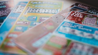 A selection of colorful lottery scratch-off tickets placed on a flat surface near a window. The image has a shallow depth of field, focusing on the tickets while blurring the background. The tickets display various games and prize amounts.
