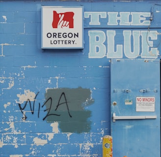 A weathered blue brick wall featuring a 'Oregon Lottery' sign with a stylized hand logo. The word 'BLUE' is painted in large letters next to a utility door with a 'No Minors Permitted' sign. There is graffiti reading 'VISA' on the wall, partially covering a green patch. An old yellow metal post and an orange traffic cone are positioned in front of the wall. A tree can be seen in the background above the wall.