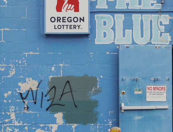 A weathered blue brick wall featuring a 'Oregon Lottery' sign with a stylized hand logo. The word 'BLUE' is painted in large letters next to a utility door with a 'No Minors Permitted' sign. There is graffiti reading 'VISA' on the wall, partially covering a green patch. An old yellow metal post and an orange traffic cone are positioned in front of the wall. A tree can be seen in the background above the wall.