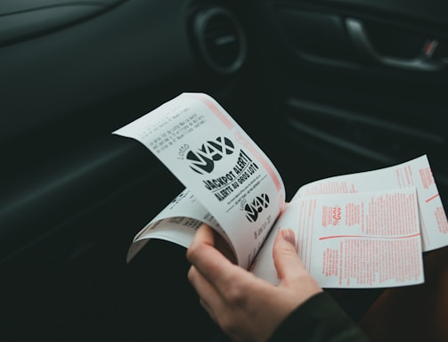 A person is holding and examining what appears to be lottery tickets inside a car. The image is focused on the tickets and the person's hands, with parts of the car interior visible in the background.