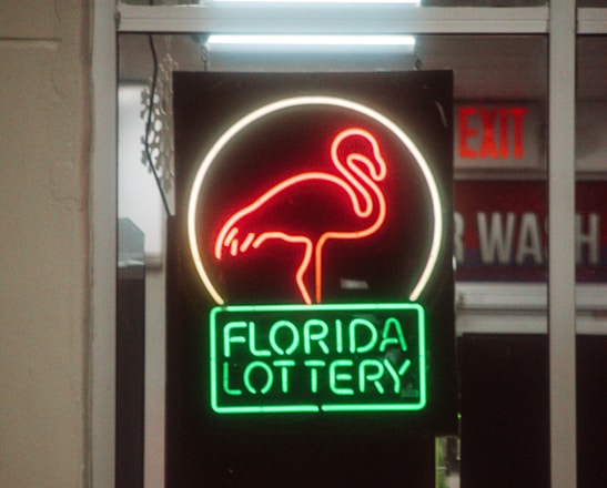 A neon sign depicting a flamingo above the words 'Florida Lottery' is prominently displayed. The sign is illuminated in bright red and green colors, and it is framed by glass panels. An 'EXIT' sign is visible in the background.
