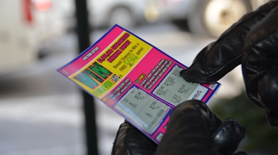 A person wearing black gloves is holding a colorful lottery scratch-off ticket. The ticket includes information about a $1,000,000 guaranteed minimum payout and features multiple games with various prize amounts.