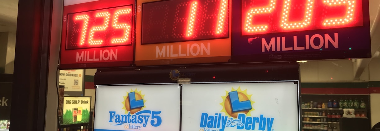 A signboard inside a convenience store displays the current jackpot amounts for various lottery games. There are bright, illuminated numbers in red and green showing the prize amounts: Powerball is at 725 million, Super Lotto Plus at 11 million, Mega Millions at 205 million, Fantasy 5 at 71 thousand, and Daily Derby at 179. In the background, shelves with various drinks and products are visible, along with promotional posters.