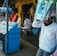 A street vendor stall displaying an array of lottery tickets clipped to a bright blue kiosk. A man in a red shirt leans on the counter, appearing thoughtful, while another man in a white shirt adjusts his hat and walks by. A variety of products hangs from strings above, and a colorful display fills the background with vibrant signs and packets. The scene suggests a busy, lively marketplace.